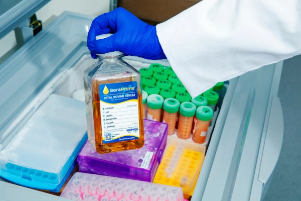 person wearing medical gear holdign up serum bottle with test tubes in background