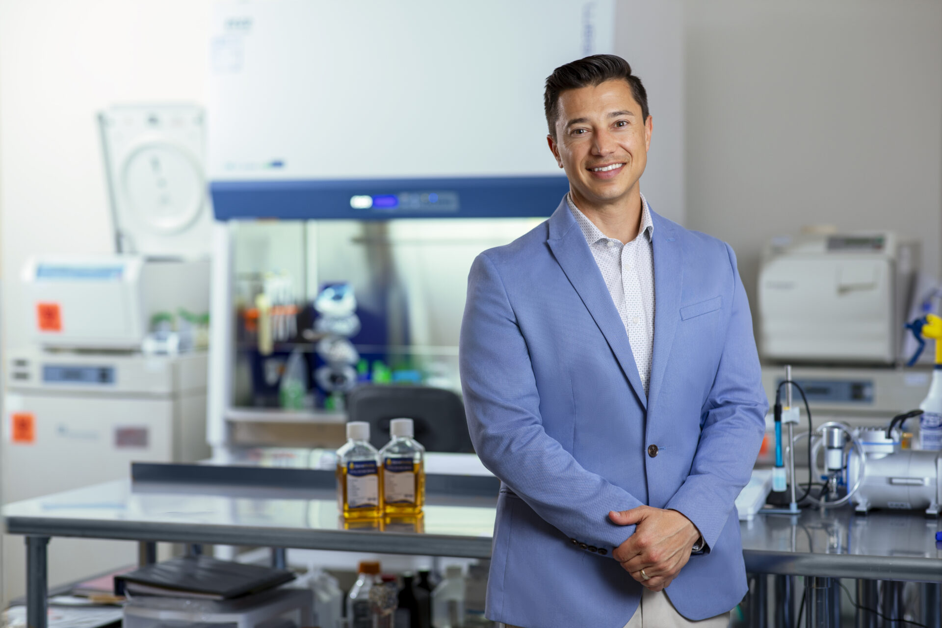 A man in a light blue blazer smiles while standing in a modern laboratory. Behind him are scientific equipment, machines, and two bottles with amber liquid on a metal table.