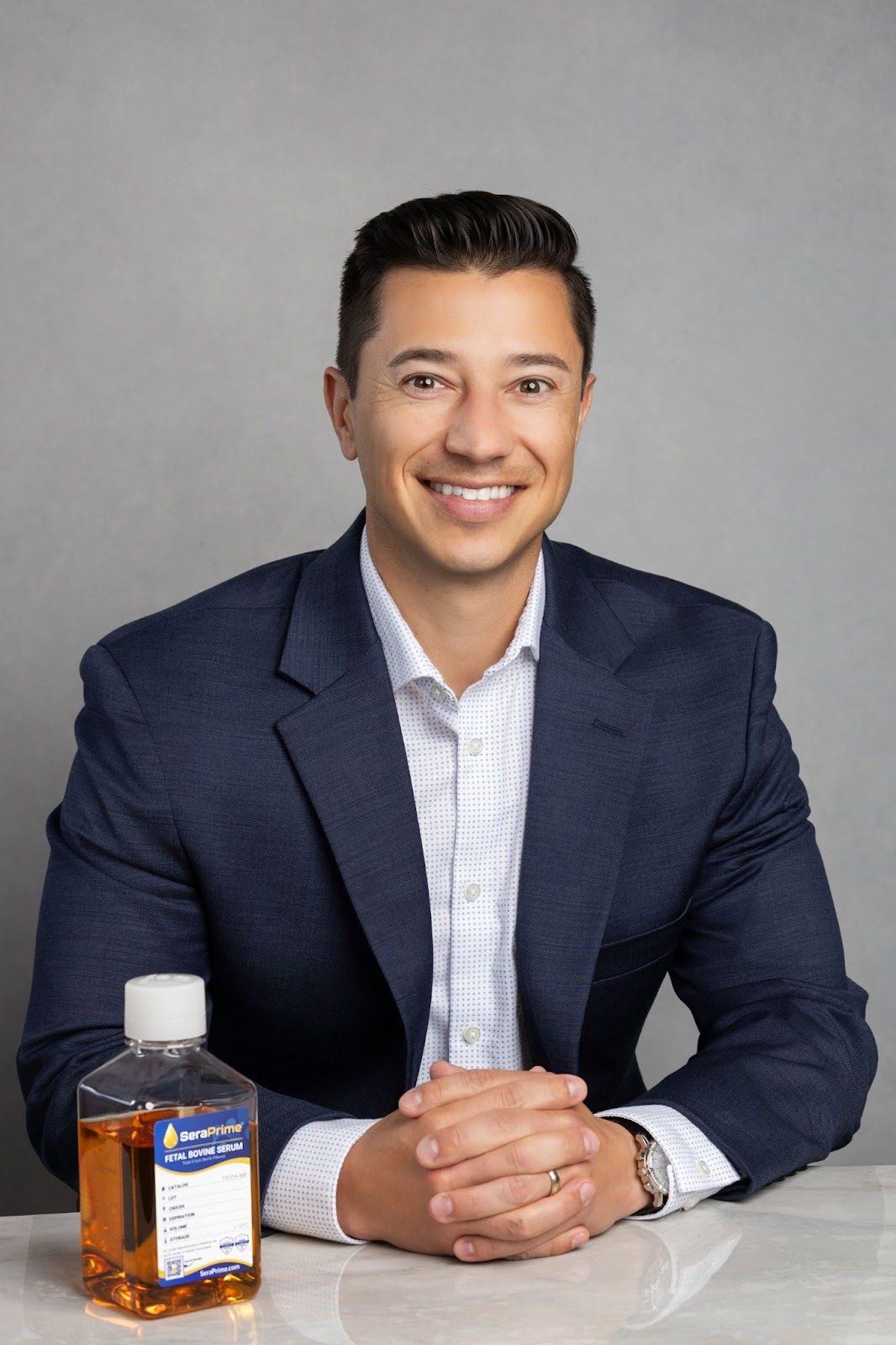A man in a dark blue suit and white shirt sits at a table, smiling with his hands clasped. In front of him is a clear bottle labeled "Seraprive," promoted as an alternative during the FBS shortage.
