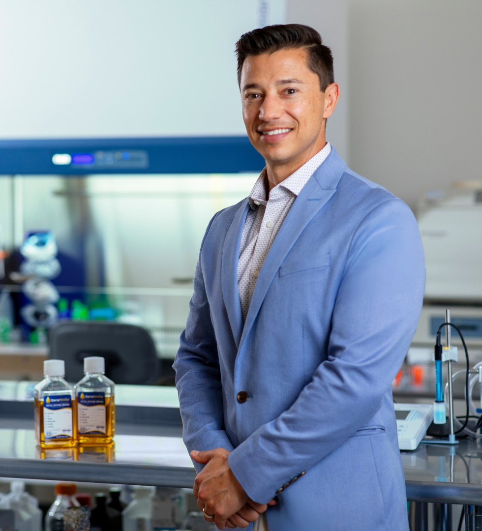 Image of Tom Kutrubes in blue blazer in lab next to 2 regular sized bottles of serum from labeled SeraPrime.