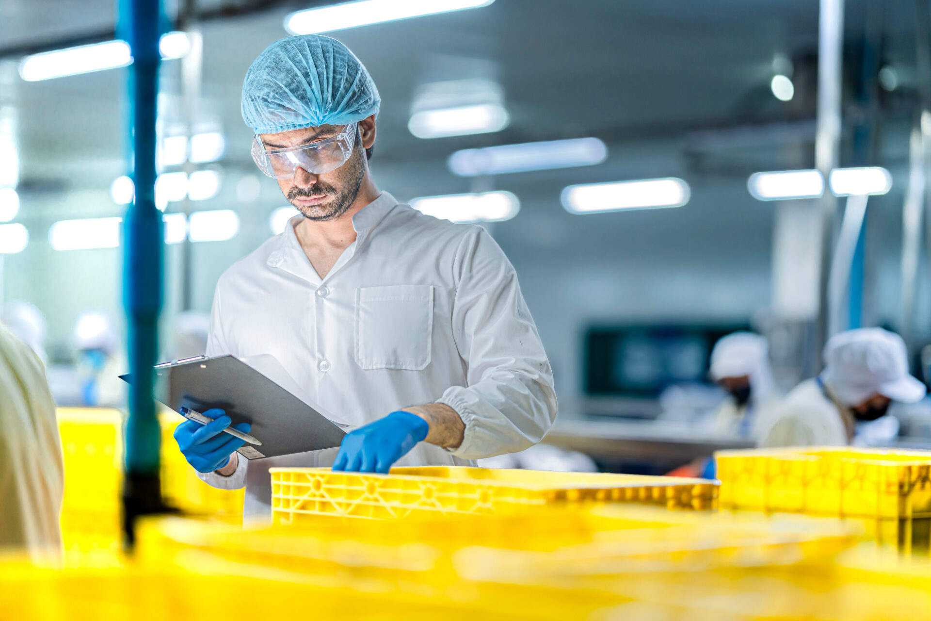 A man in protective clothing, gloves, hairnet, and safety glasses inspects yellow crates in a brightly lit factory, holding a clipboard—demonstrating vigilance against lab supply chain risk. Other workers in similar attire are visible in the background.