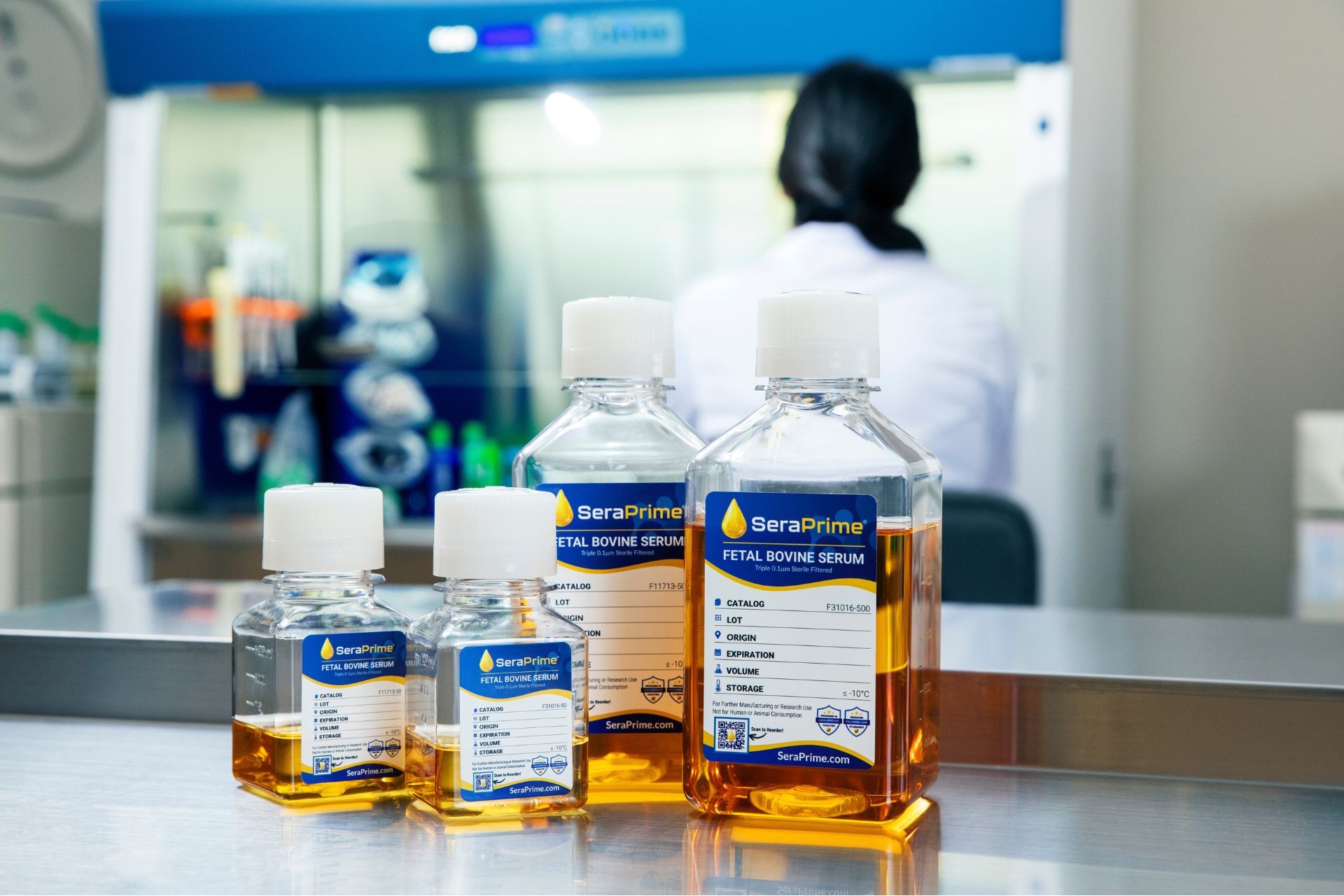 Four bottles of SeraPrime Fetal Bovine Serum with orange liquid are on a lab bench. In the background, a person in a lab coat works in front of a biosafety cabinet with blurred lab equipment visible. Four bottles of SeraPrime Fetal Bovine Serum with orange liquid are on a lab bench. In the background, a person in a lab coat works in front of a biosafety cabinet with blurred lab equipment visible.